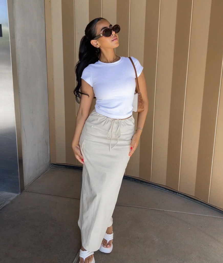 Woman in a chic summer outfit featuring a white cropped tee and beige maxi skirt, accessorized with sunglasses and a shoulder bag.